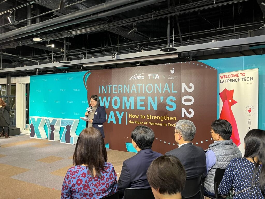 慶萱拿著麥克風在台前主持，背後有法國科技在台灣（La French Tech Taiwan）2023年國際婦女節活動的橫幅