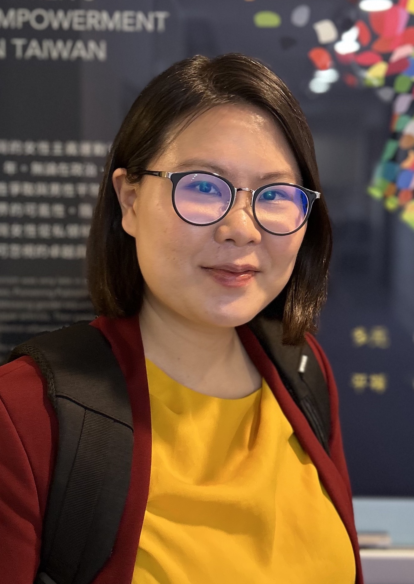 慶萱微笑看向鏡頭，她是一名台灣女性，戴著眼鏡、身穿暗紅色的西裝外套與黃色上衣